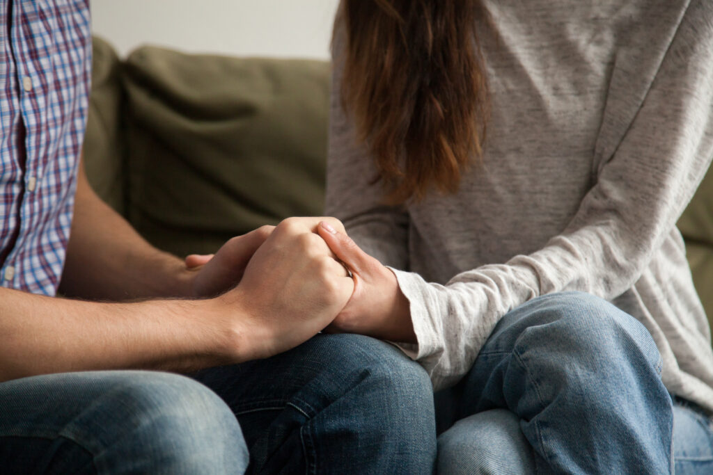 Image shows close-up of a man and a woman sitting on a couch, holding hands. You can't see their faces, their clasped hands are the focus. 